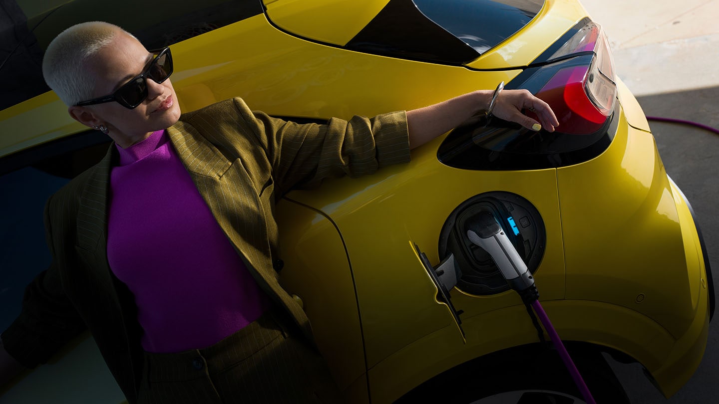 Une femme s'appuyant sur un Ford Capri électrique jaune connecté à un chargeur.